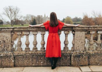 woman in red dress looking at body of water and trees during daytime